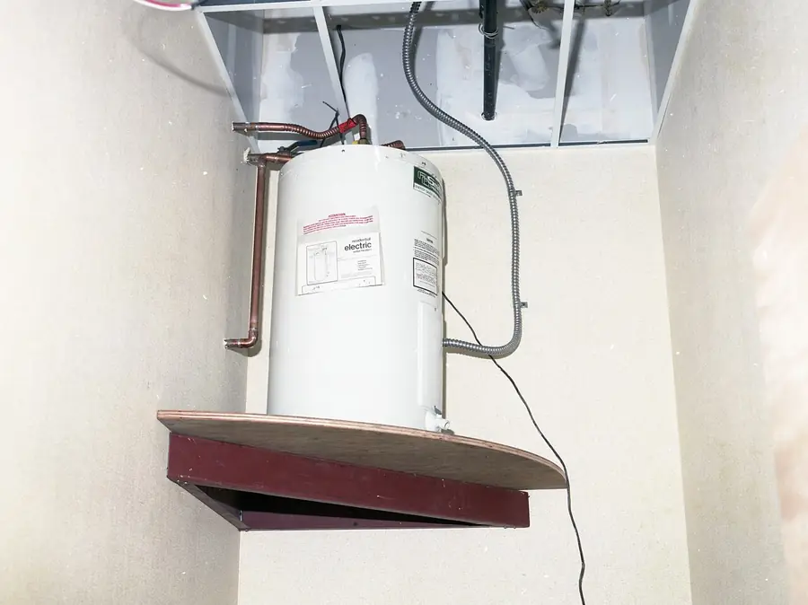 Small tank water heater hung on a wall with piping and a shelf below