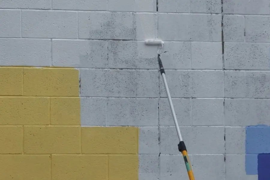 Paint roller on a long pole rolling white paint over a block wall