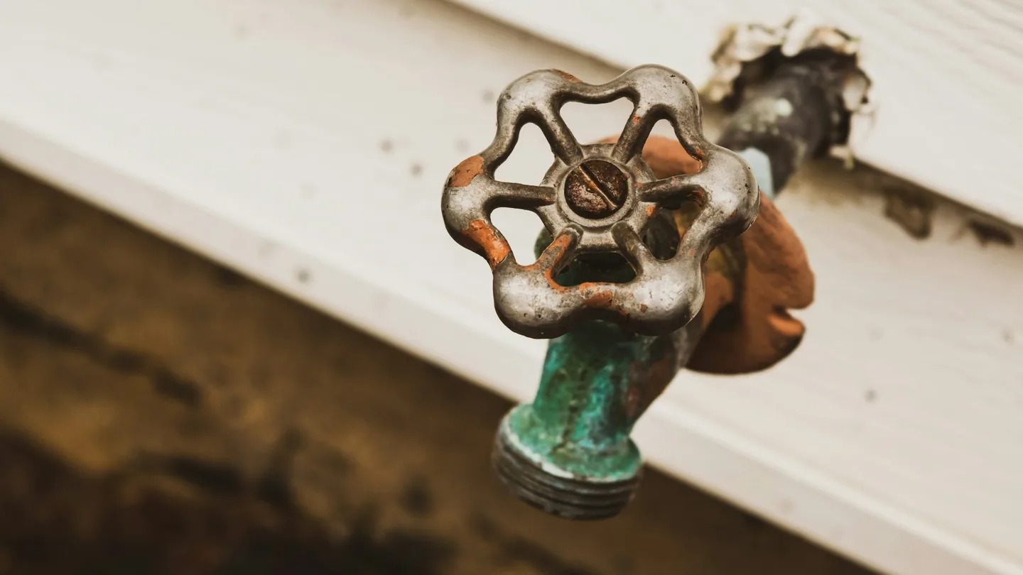 Worn outdoor hose bib with a round metal handle, coming out of white house siding
