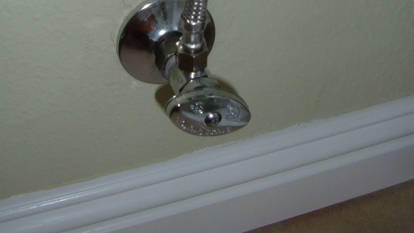 Chrome angle stop valve on a wall above the baseboard, with a braided supply line