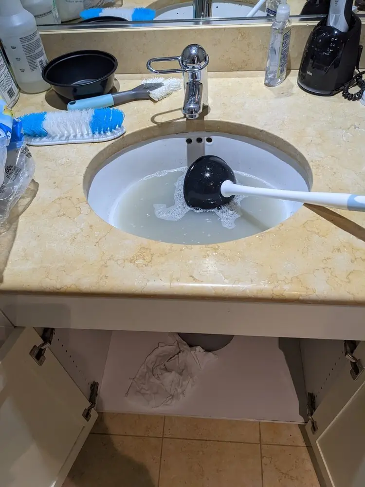 Bathroom sink drain being cleared with a small cup plunger and brush